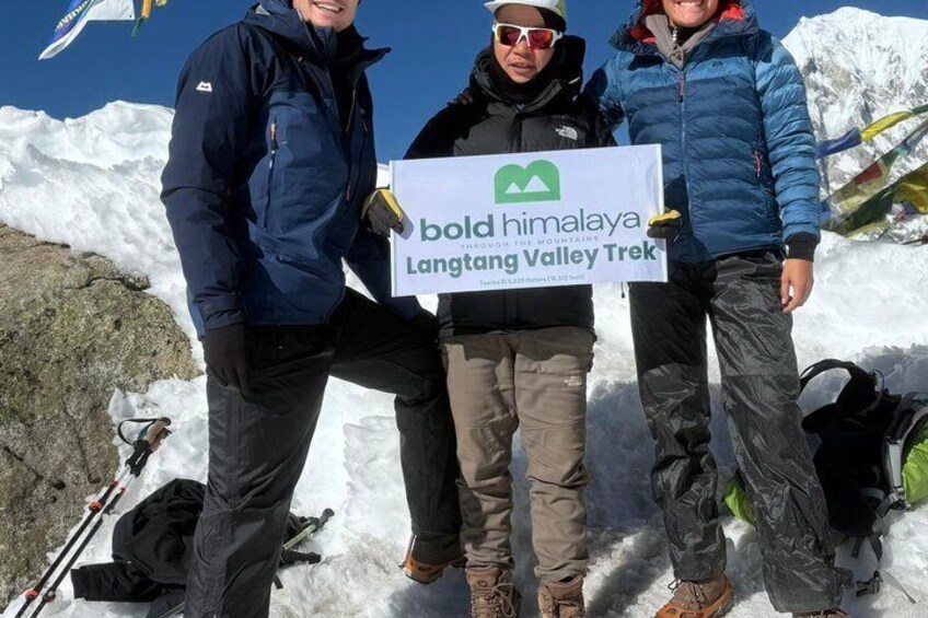 Three people standing on a snowy mountain with a sign that reads 'bold himalaya Langtang Valley Trek.' 1