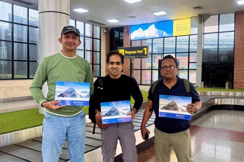 Three people standing in an airport baggage claim area with a sign indicating 'Baggage Belt 1'. 2