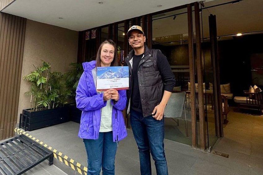 Two people holding a certificate outside a building with glass doors and a bench in the foreground. 3