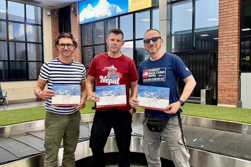 Three people holding certificates in front of a building with a mountain image on the wall. 5