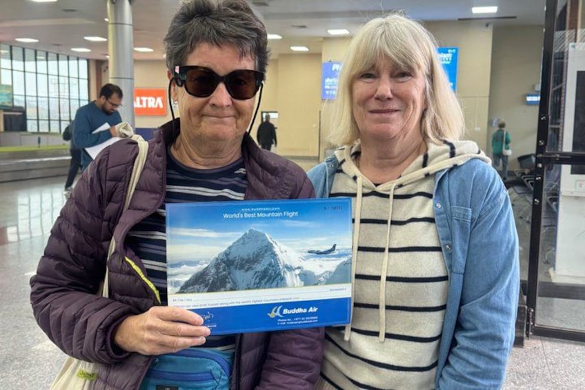 Two people holding a brochure with a mountain image, standing in an airport with visible signage and a glass wall. 5