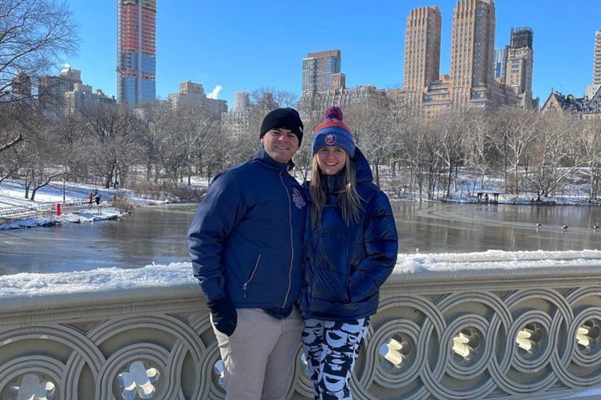 Two people standing in a park with a river and city skyline in the background. 1