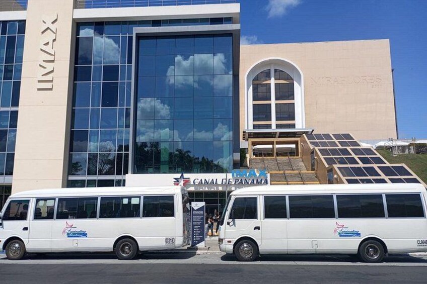 A white bus with 'CANADLE PANAMA' written on it parked in front of a building with a large glass window and a sign that reads 'IMAX'. 5