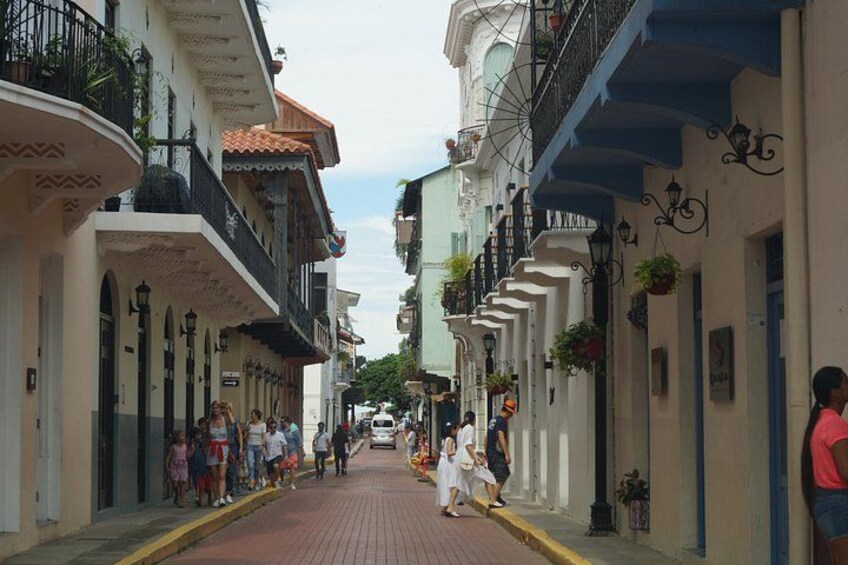 A narrow street lined with buildings, some with balconies and hanging plants. 2