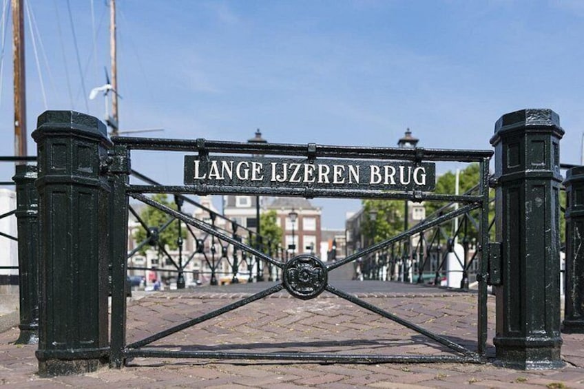 En metalport med inskriften 'LANGE IJZEREN BRUG' i forgrunden, med en brostensbelagt gade og bygninger i baggrunden. 2
