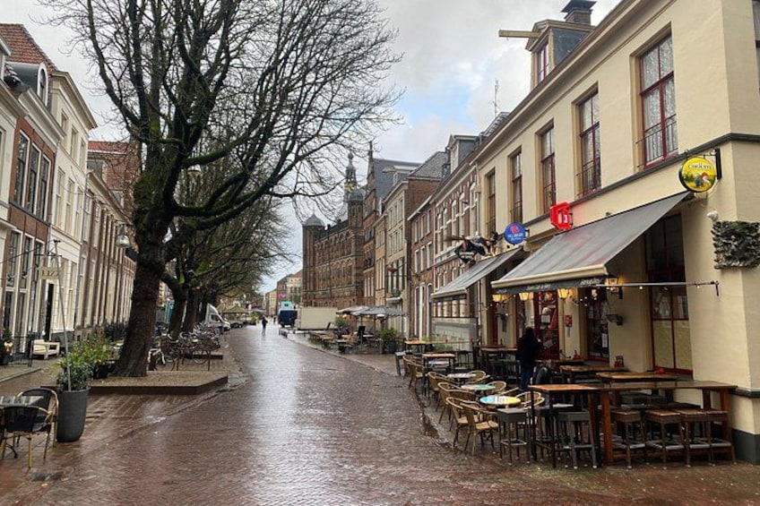 A wet cobblestone street lined with buildings, outdoor seating at a café, and a large tree. 4