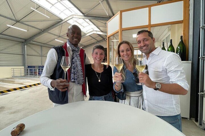 Four people holding wine glasses in a warehouse with wine bottles in the background. 3