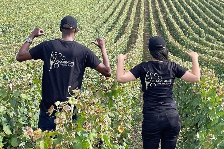 Two people in a vineyard wearing black t-shirts with 'Champagne Dream Tour' printed on the back. 5