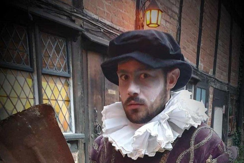 A person in historical costume with a ruffled collar and a hat, standing in front of a building with a window and a lantern. 1