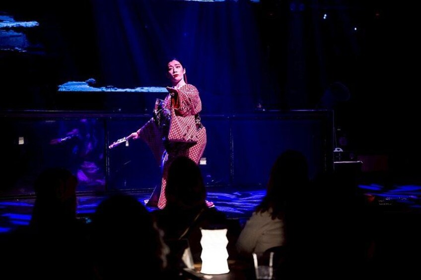 Une personne se produisant sur scène avec un microphone et une guitare, un public au premier plan et un fond sombre éclairé par des projecteurs. 3
