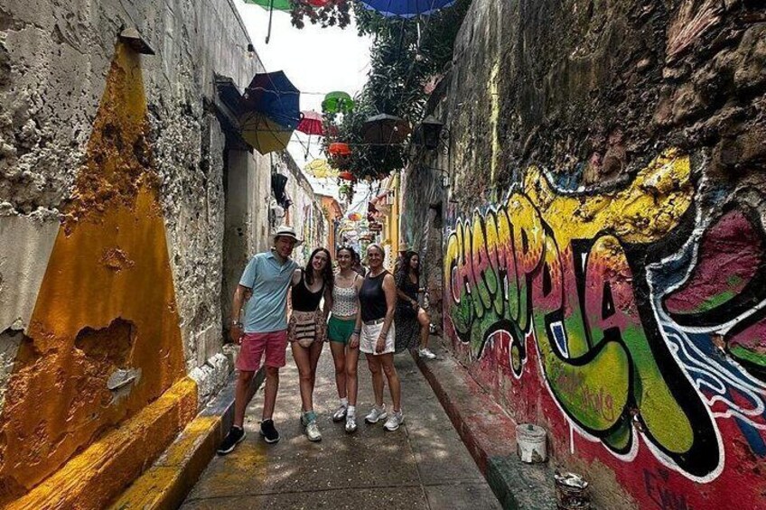 Une ruelle étroite dont les murs sont couverts de graffitis, un groupe de personnes qui se tiennent debout, et un mur jaune et orange sur la gauche. 1