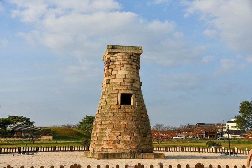 A stone tower with a small window, surrounded by a fence and a landscaped area with trees and a traditional building in the background. 2