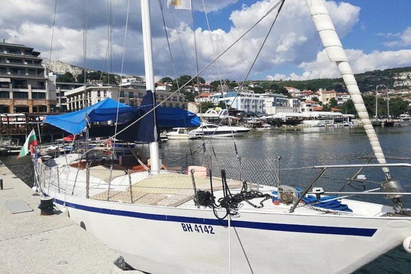 A sailboat docked at a marina with buildings and hills in the background. 1