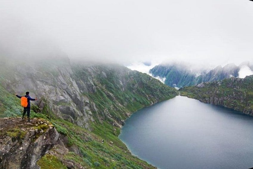 A hiker on a mountain ridge overlooking a calm lake with steep, green slopes and misty mountains in the background. 3