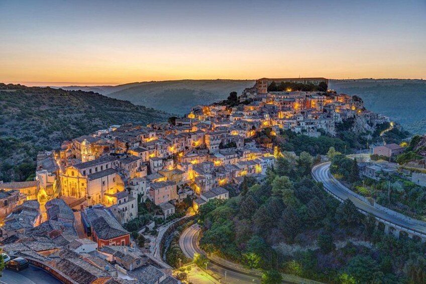 A coastal town at dusk with illuminated buildings, a prominent fort, and winding roads. 1