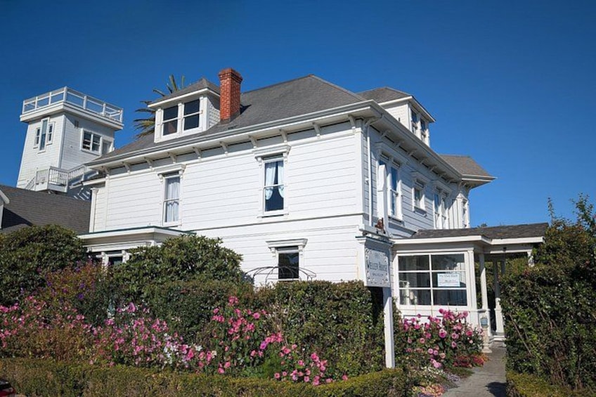 A white two-story house with a red chimney, surrounded by pink flowers and greenery. 2