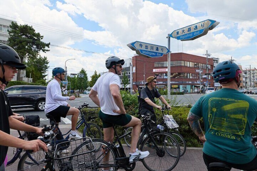 A group of cyclists on a city street with directional signs and buildings in the background. 3