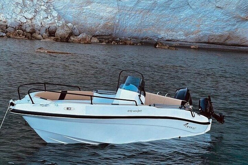 A small white motorboat with a cabin and seating area, floating on calm water near a rocky shoreline. 2