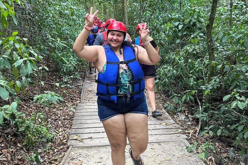 A person wearing a blue life jacket and a red helmet, standing on a wooden boardwalk surrounded by dense green foliage. 4