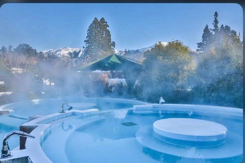 A hot spring with steam rising, surrounded by snow-covered mountains and trees. 2