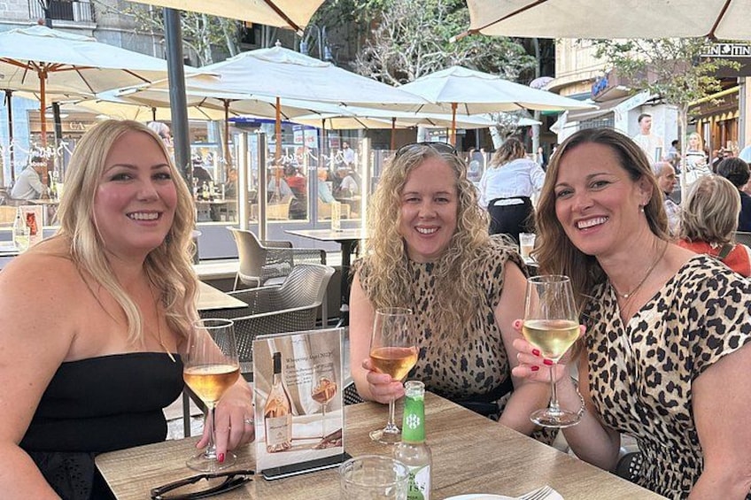 Three women sitting at an outdoor table with drinks, surrounded by umbrellas and other diners. 2