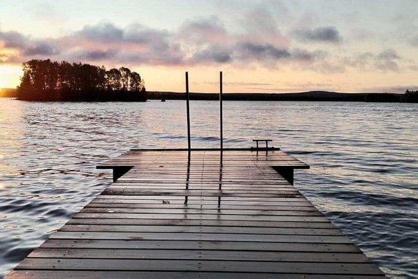 En træbrygge der strækker sig ud i rolige vand, med en solnedgang i baggrunden. 2