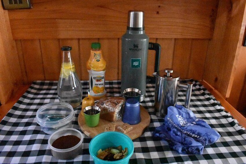 Una mesa a cuadros con una botella de jugo de naranja, un termo, una cafetera, una bolsa de aperitivos y un paño azul. 2