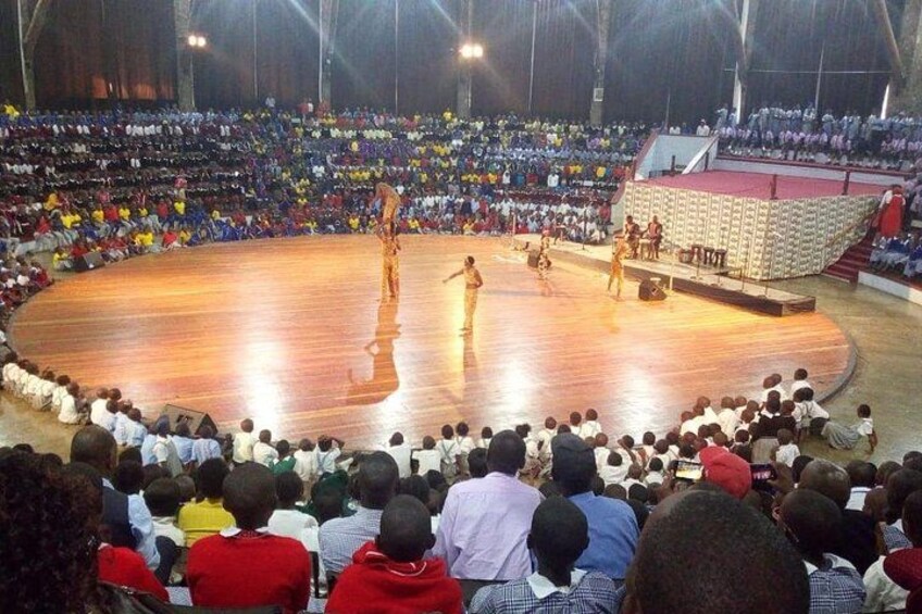 A large indoor arena with a wooden stage, an audience seated in rows, and performers on stage. 3
