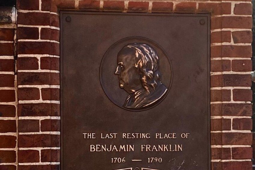 A plaque with a bust and text stating 'The Last Resting Place of Benjamin Franklin 1706-1790' mounted on a brick wall. 3