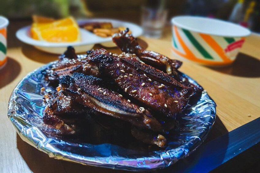 A plate of barbecued ribs with a foil cover, accompanied by a side dish and a beverage in the background. 4