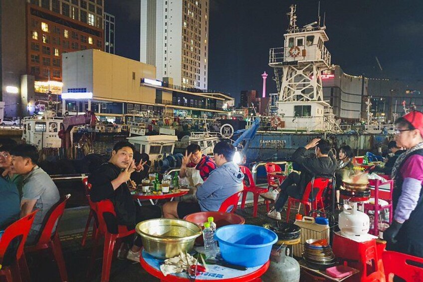 A bustling waterfront dining area with people seated at tables, boats docked in the background, and a prominent tower structure. 5