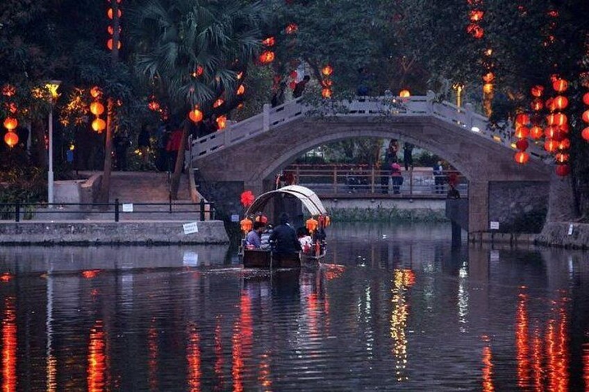 Een brug over het water, met weerspiegelingen van lichtjes, palmbomen en een boot met passagiers. 1