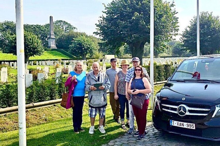 A group of people standing in a cemetery with a monument in the background and a car parked nearby. 1