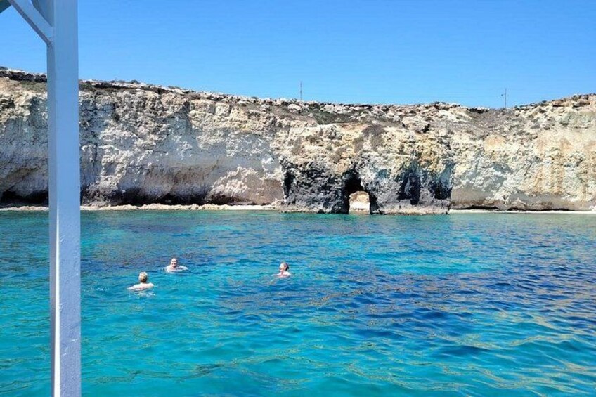 A clear blue sea with three people swimming, a rocky cliff, and a cave entrance. 2