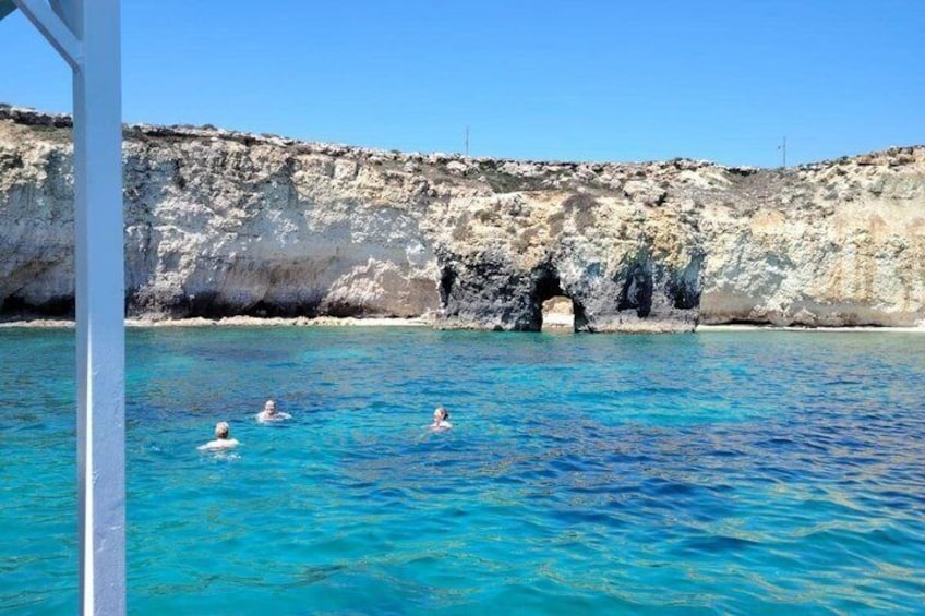 A clear blue sea with three people swimming, a rocky cliff, and a cave entrance. 2