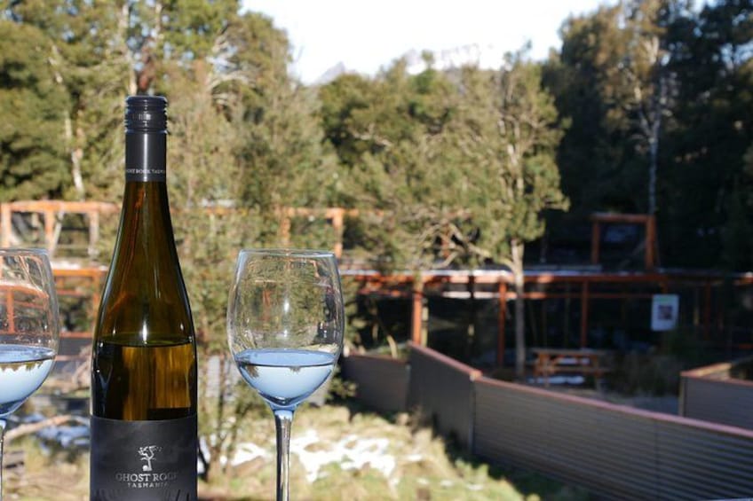 A bottle of Ghost Rock wine and two glasses of wine on a table with a view of a forest and a wooden structure in the background. 3