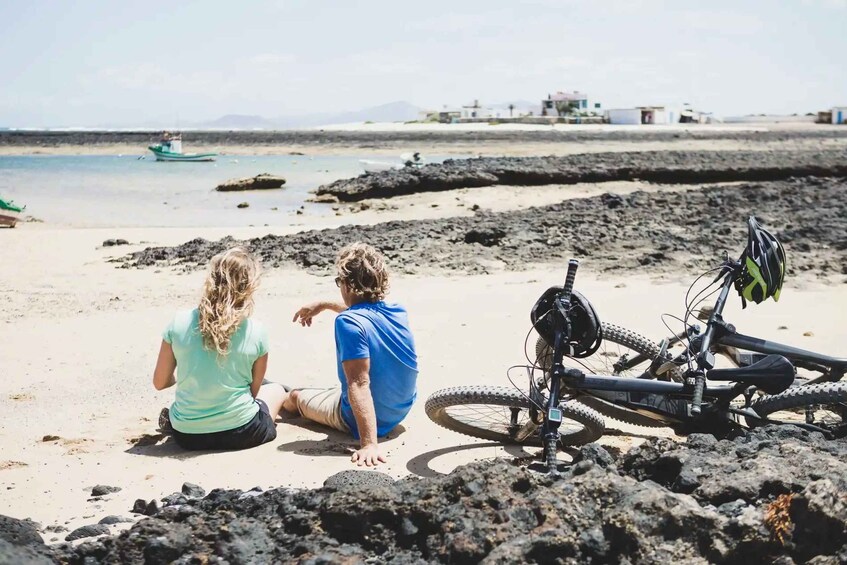 Two people sitting on a beach with a bicycle and a boat in the background. 5