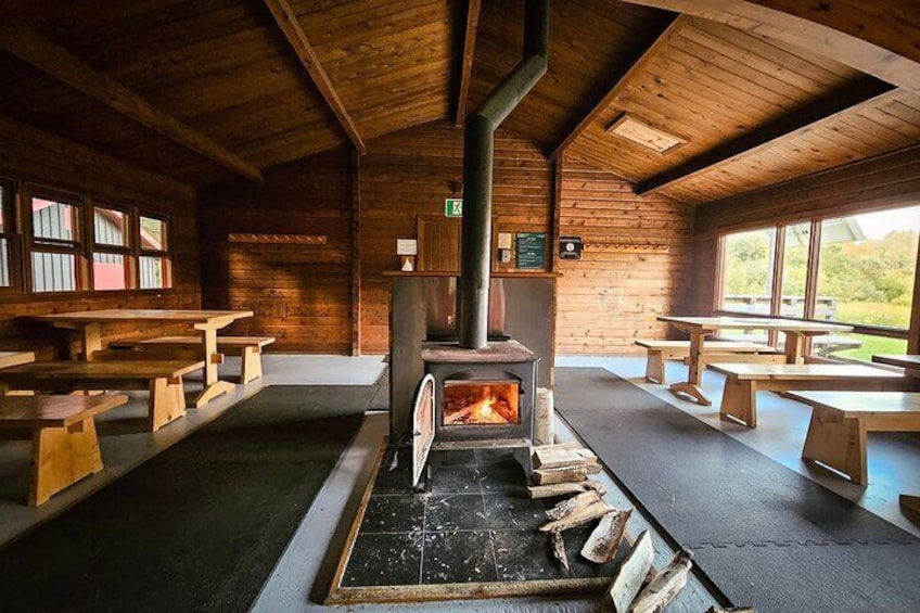 A rustic wooden cabin with a central wood-burning stove, benches, and large windows. 5