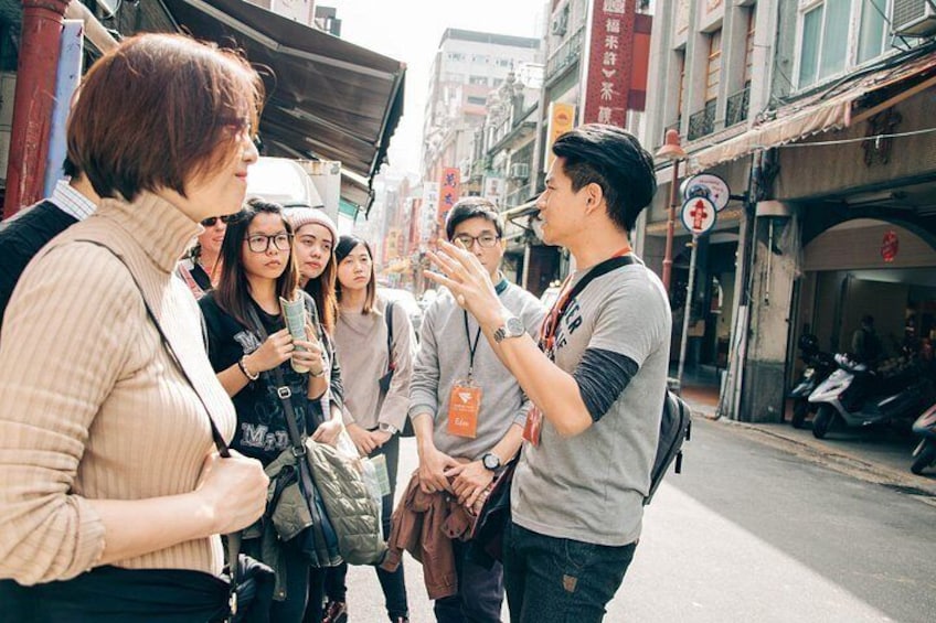 A group of people on a city street, some holding drinks, with buildings and a signboard in the background. 2
