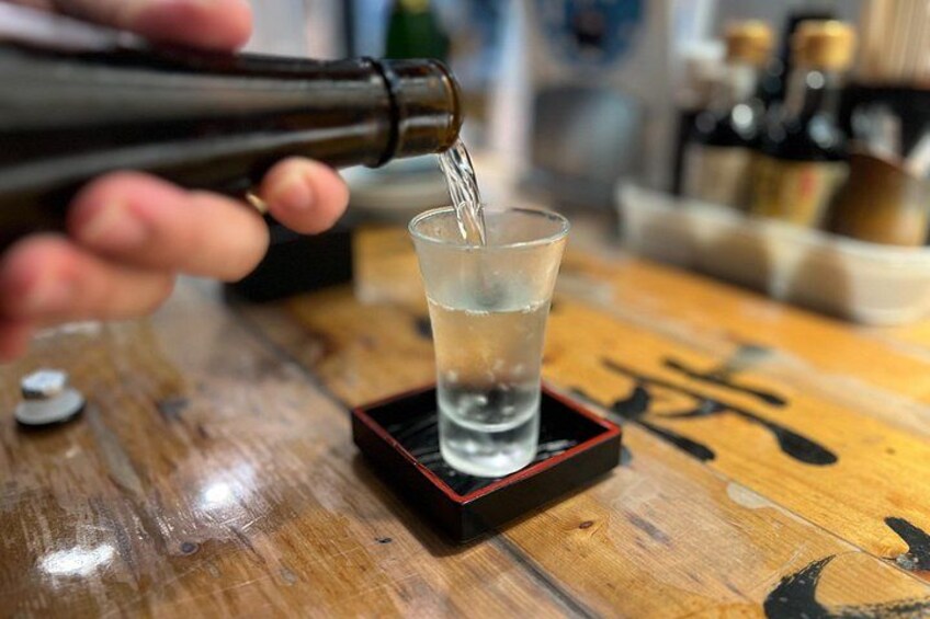 En hand häller en klar vätska från en flaska i ett litet glas som står på ett träbord. 3