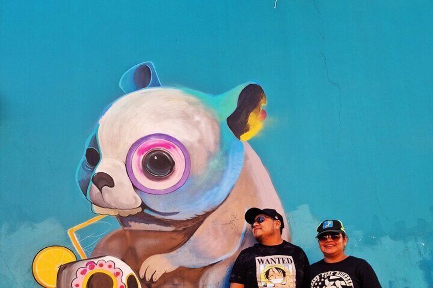 Ein Wandgemälde mit einem großen Panda mit einem rosa Auge, einer Person in einem 'WANTED'-Shirt und einer weiteren Person mit Mütze. 1