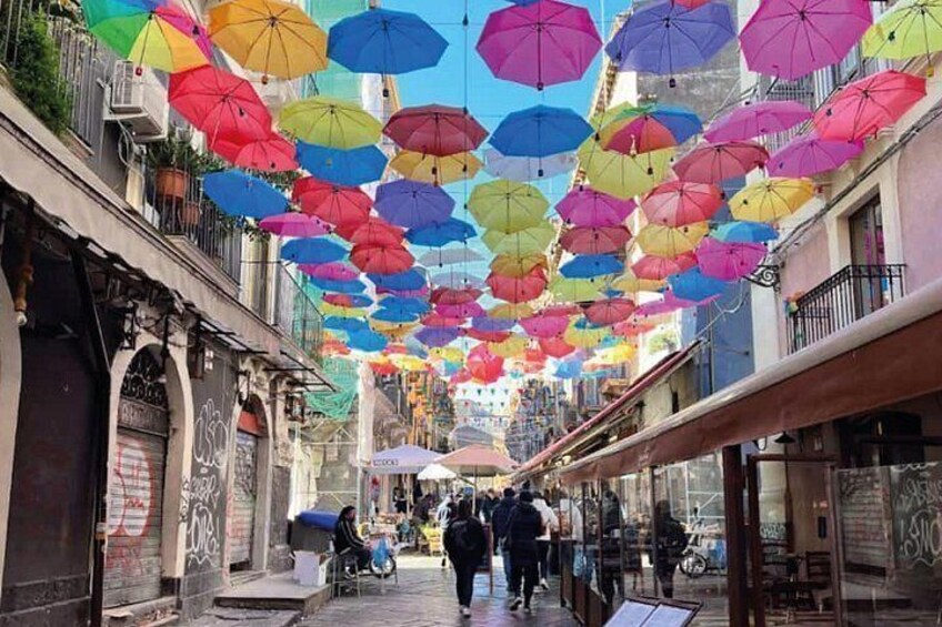 A street with colorful umbrellas hanging overhead, shops on either side, and people walking. 4