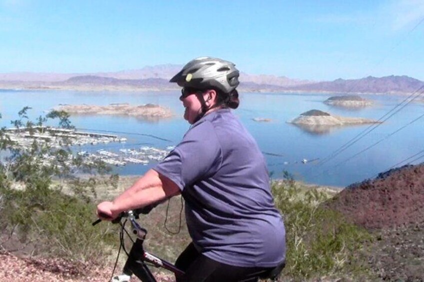 A person riding a bicycle on a trail with a scenic view of a lake and mountains in the background. 1