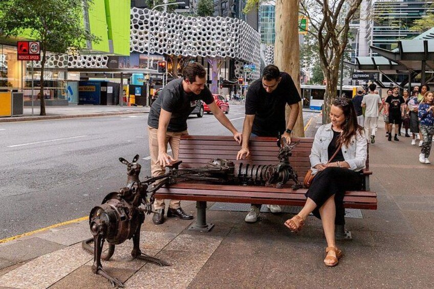 Un banc orné d’une sculpture représentant un animal mécanique, une personne debout à côté, et une autre personne assise sur le banc. 5