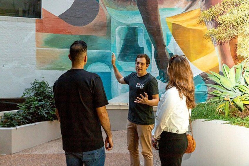 Trois personnes se tiennent devant une fresque colorée, ornée de formes abstraites, avec un pot de fleurs placé à droite. 3