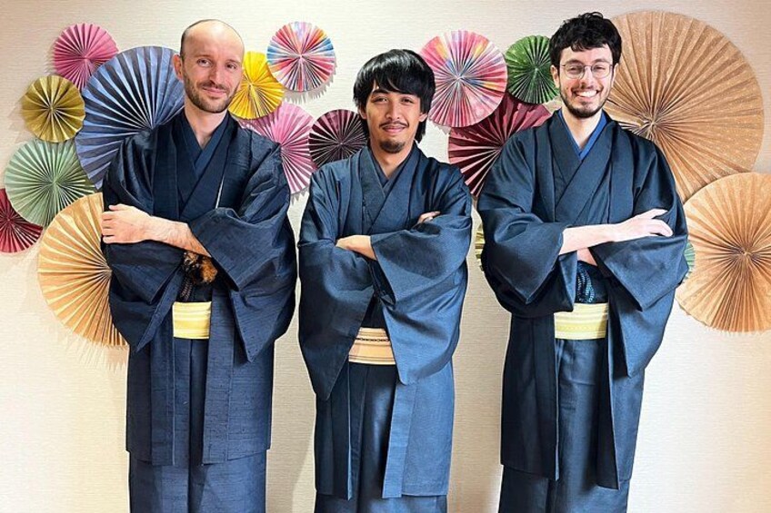Trois personnes, vêtues d’un kimono bleu traditionnel, les bras croisés, se tiennent devant un mur décoré d’éventails en papier aux couleurs vives. 5