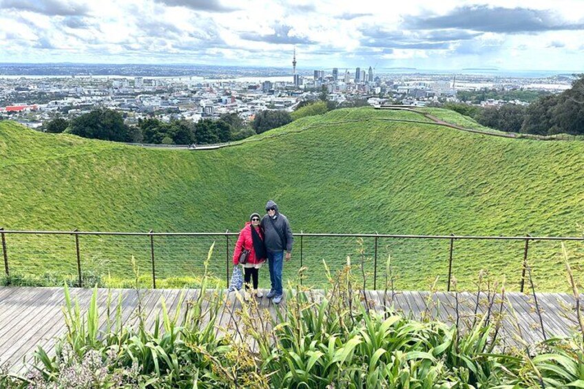 A couple standing on a wooden boardwalk overlooking a cityscape with a hill in the foreground. 4