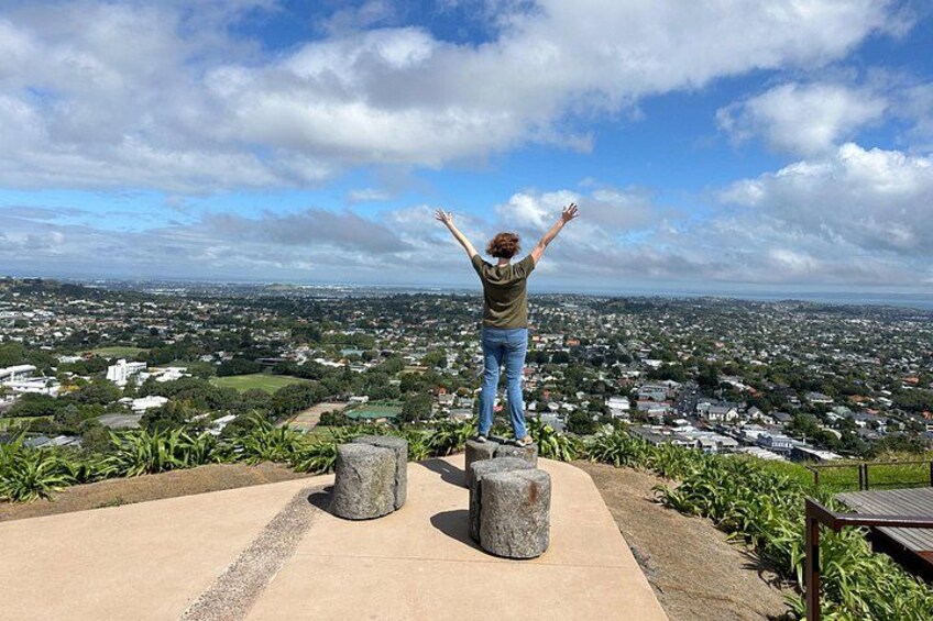 A person standing on a concrete platform with arms raised, overlooking a cityscape with houses and greenery. 2