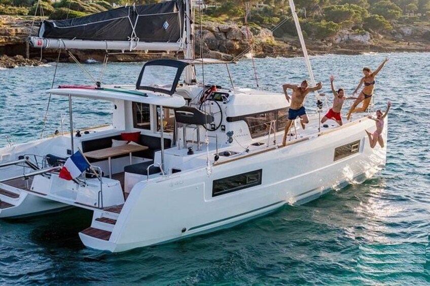 En vit yacht med en flagga, människor som hoppar från fördäcket, stilla vatten och en klippig kustlinje i bakgrunden. 1