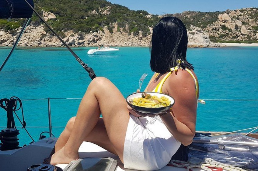 A person sitting on a boat, holding a plate of food, with a rocky coastline and clear blue water in the background. 3